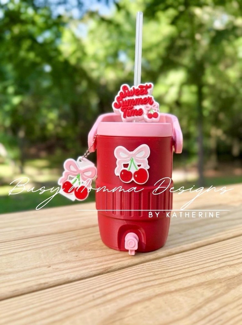 May include: Red insulated cooler with a pink lid and handle, decorated with cherry and bow decals. A straw protrudes from the top, and a cherry charm hangs from the side. The words "Sweet Summer Time" are on a topper.