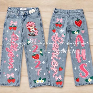 May include: Light blue denim jeans featuring hand-painted designs including a cartoon character, strawberries, pink bows, and white flowers. The text reads "Allyson", "2027", "Senior", and "AHS" in pink and white script and block lettering.
