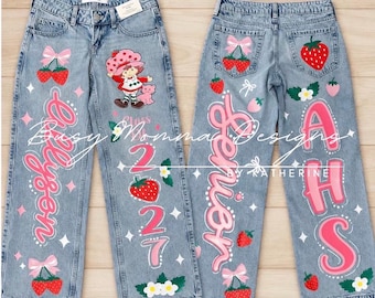 Può includere: Jeans in denim azzurro con disegni dipinti a mano che includono un personaggio dei cartoni animati, fragole, fiocchi rosa e il testo "Allyson", "2027", "Senior" e "AHS". I jeans presentano una classica struttura a cinque tasche con cerniera.