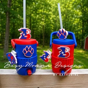 May include: Two red and blue insulated water coolers with straws. Each cooler has a matching lid, handle, and decorative charm. One has a paw print and the other has a letter "B" design. The text "Busy Momma Designs" and "By Katherine" are visible.