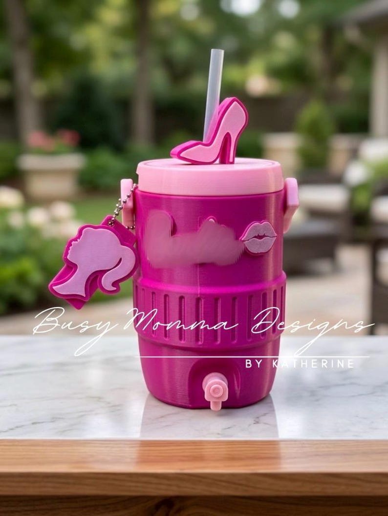 May include: A pink and fuchsia water cooler with a high-heeled shoe accent on the lid. The cooler has a straw, a charm with a silhouette, and the text "Busy Momma Designs BY KATHERINE".