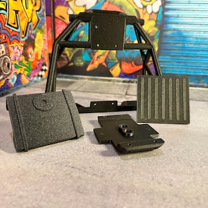 May include: A collection of black, textured, 3D-printed model parts. The components include a roll cage, a rectangular panel with vertical lines, a square panel, and a small, irregularly shaped piece. The parts are arranged on a light gray surface.