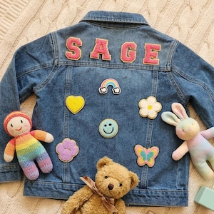 Puede incluir: Chaqueta vaquera azul con el nombre "SAGE" en letras rosas en la espalda. La chaqueta está decorada con parches que incluyen un arcoíris, un corazón, una flor, una carita sonriente, una huella de pata y una mariposa. Una muñeca de punto, un oso de peluche y un conejito de peluche también están en la imagen.