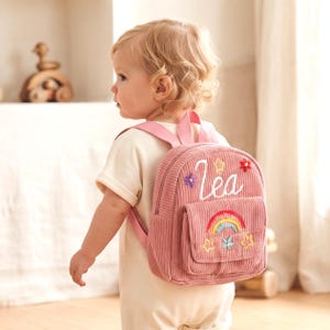 Puede incluir: Una mochila de pana rosa con el nombre Lea bordado en letra blanca, decorada con flores, estrellas y un arcoíris de colores en el bolsillo delantero. La mochila incluye correas rosas a juego y un compartimento principal con cremallera.