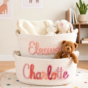 Peut inclure: Deux paniers en corde blanche superposés, avec les noms "Eleanor" et "Charlotte" en script rose et orange. Le panier supérieur contient une couverture blanche et deux lapins en peluche. Le panier inférieur contient un ours en peluche.