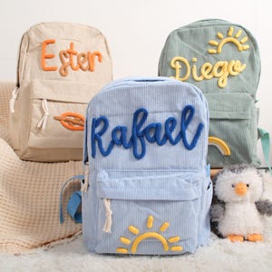 Puede incluir: Tres mochilas de pana en beige, azul claro y verde salvia. Cada mochila presenta un nombre bordado en un color contrastante: "Ester" en naranja, "Rafael" en azul y "Diego" en amarillo. La mochila azul claro tiene un diseño de sol.