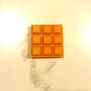 May include: A bright orange square with a grid of nine raised squares. The object has a textured surface, possibly 3D-printed. The design is simple and geometric, with a uniform colour throughout.