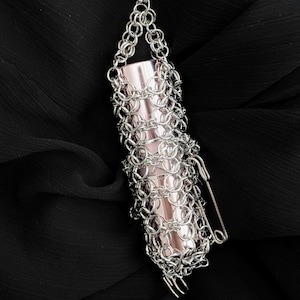 May include: A silver chainmail lipstick holder containing a pink lipstick. The lipstick is secured with a safety pin. The holder hangs from a delicate chain against a black fabric backdrop.