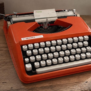 May include: A bright orange Hermes Baby portable typewriter with a white keyboard featuring black lettering. A piece of paper with the text bonus is inserted into the carriage. The machine has a metal frame and a standard QWERTY-style layout.