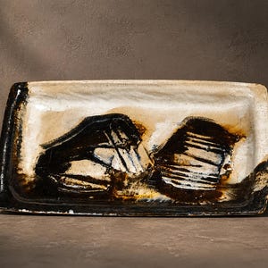 Op de afbeelding: Rechthoekige keramische schaal met een crèmekleurig oppervlak en abstracte zwarte en bruine penseelstreken. De schaal heeft een donkerbruine rand en is geschikt om te serveren of tentoon te stellen.