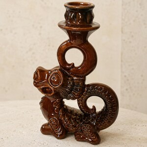 Pode incluir: Um castiçal de cerâmica castanho brilhante em forma de dragão. A criatura é representada numa posição sentada com uma cauda enrolada e escamas texturizadas. Um copo circular para velas assenta sobre a cabeça do dragão.