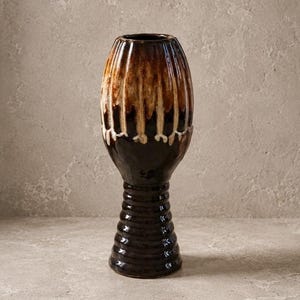 May include: A tall ceramic vase with a glossy finish, featuring a dark brown base and a ribbed, tapered stem. The upper section is bulbous with vertical fluting, showcasing a drip glaze effect that transitions from dark brown to cream and tan.