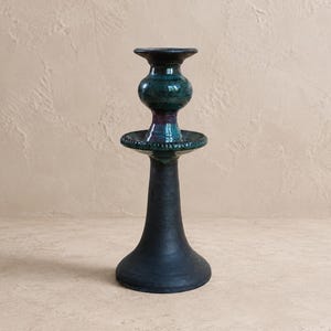 May include: A ceramic candlestick holder featuring a matte black flared base and a glossy, multi-toned upper section. The top portion displays a bulbous shape with a deep teal and dark purple glaze, finished with a wide, circular drip tray.