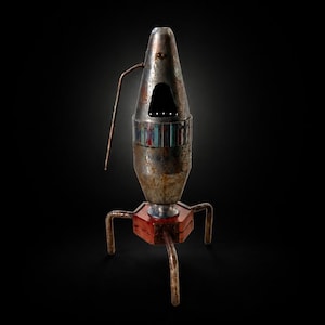 May include: A metallic rocket-shaped sculpture with a weathered, rusted finish. It features a central triangular opening, a band of vertical multicolored stripes, a single thin protruding antenna, and a red hexagonal base with three curved metal legs.