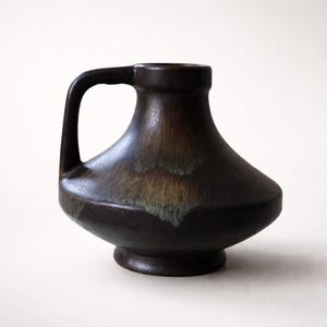 May include: A dark brown ceramic vase with a handle. The vase has a wide base and a flared top, with a handle on one side. The glaze has a mottled appearance with hints of green and brown.