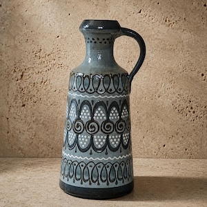 Può includere: Una brocca in ceramica con corpo affusolato e un manico singolo. La superficie presenta uno smalto grigio-azzurro chiaro decorato con motivi dipinti a mano in marrone scuro e bianco, tra cui archi, spirali e punti ripetuti sull'esterno.