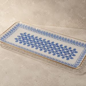 Op de afbeelding: Een rechthoekig glazen serveerblad met een witte basis en een herhalend blauw bloemmotief in het midden en langs de randen. Het blad is afgewerkt met een dunne goudkleurige metalen rand die de gehele omtrek van het object omlijnt.