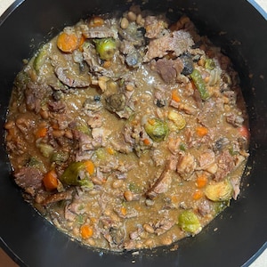 May include: A top-down view of a black pot filled with a hearty stew. The stew contains chunks of meat, white beans, orange carrots, green brussel sprouts, and mushrooms in a brown gravy.