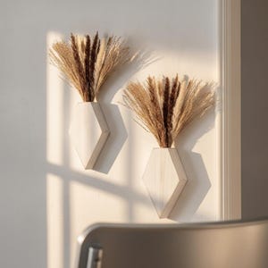 May include: Two white geometric wall-mounted vases, each holding a dried floral arrangement of beige and dark brown pampas grass and plumes. The vases have a faceted, diamond-like shape and are mounted side-by-side on a plain white wall.