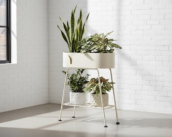 Puede incluir: Un soporte para plantas de dos niveles de color crema, con una parte superior rectangular y un estante inferior. El soporte contiene varias plantas en macetas con hojas verdes y abigarradas. El soporte es de metal y tiene un diseño minimalista.