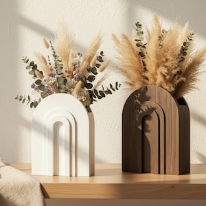 May include: Two decorative vases with arch designs, one white and one brown, each filled with dried flowers and pampas grass. The vases sit on a wooden surface, with a neutral-toned background and soft lighting.