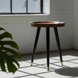 May include: A small, round side table with a dark wood top and black legs. The legs have gold-colored tips. The table is set against a white brick wall and a window, with a large green leaf in the foreground.