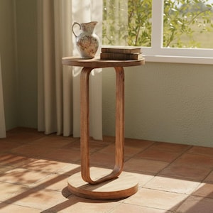 May include: A wooden side table with a round top and a unique curved leg design. The table holds a decorative pitcher with floral patterns and two books. The table is set near a window with natural light.