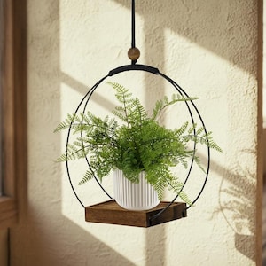 May include: A hanging planter featuring a black metal frame with a circular design. A small, square wooden shelf holds a white, ribbed ceramic pot filled with green fern plants. The planter hangs from a black cord with a wooden bead.