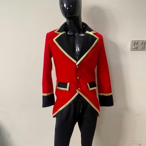 May include: A red jacket with black lapels, black cuffs, and gold trim, featuring three gold buttons down the front and two flap pockets. The jacket is paired with black trousers, displayed on a glossy black mannequin torso.