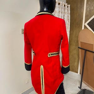 May include: A bright red tailcoat jacket featuring black cuffs and a black collar. The garment is accented with gold piping along the collar, the back waist pockets, and the central back vent. The jacket has a long, structured tail design.
