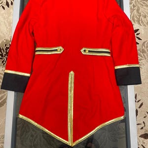 May include: A bright red tailcoat jacket featuring black cuffs and a black collar. The garment is accented with gold trim along the hem, the back vent, and the two horizontal back pockets, which are each finished with a single gold button.