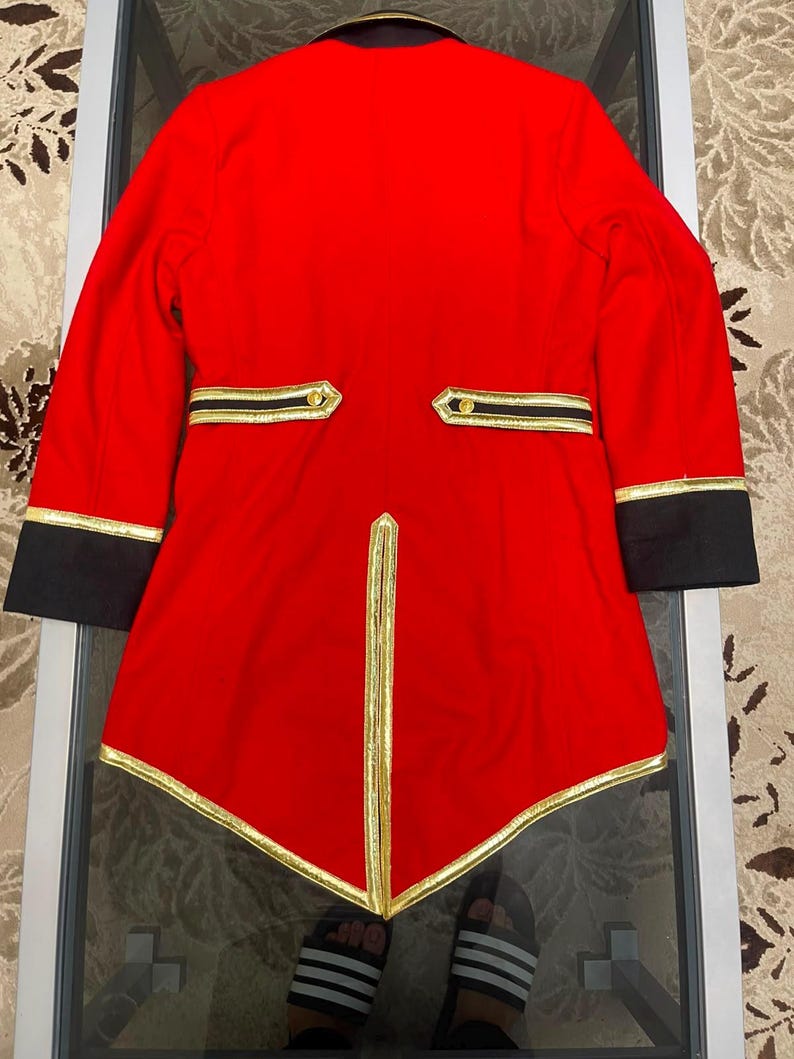 May include: A bright red tailcoat jacket featuring black cuffs and a black collar. The garment is accented with gold trim along the hem, the back vent, and the two horizontal back pockets, which are each finished with a single gold button.