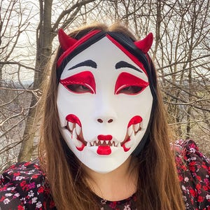 May include: A white mask with red horns, eyes, and a mouth with sharp teeth. The mask has a black and red headband. The person is wearing a floral print top. The background features bare trees.