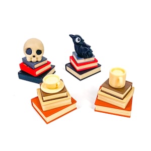 May include: Miniature figurines of a skull, a crow, a teacup and saucer, and a mug, each atop a stack of books. The books have red, orange, and blue covers. The skull is beige, the crow is black, and the teacup and mug are yellow.