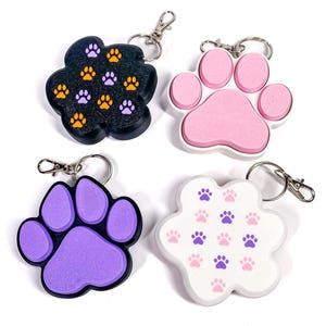 May include: Four paw-shaped keychains with metal lobster clasps. One is black with orange and purple paw prints, one is solid pink with a white border, one is solid purple with a black border, and one is white with small pink and purple paw prints.
