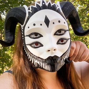 May include: A white mask featuring four eyes with black iris details, a black diamond-shaped forehead marking, and large, curved black horns. The lower portion of the mask displays intricate black and white line patterns around the mouth area.