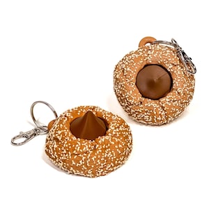 Può includere: Due portachiavi marroni a forma di ciambella con semi di sesamo bianchi e un centro marrone scuro simile al cioccolato. Ogni portachiavi ha un fermaglio e un anello color argento per attaccarlo alle chiavi o a una borsa. Su sfondo bianco.