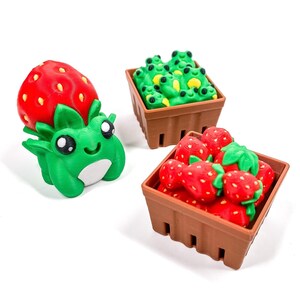 Könnte beinhalten: Eine Sammlung von 3D-gedruckten Figuren. Ein grüner Frosch mit Erdbeerkopf steht neben zwei braunen Körben. Ein Korb ist mit grünen Froschfiguren gefüllt, der andere mit roten Erdbeeren.