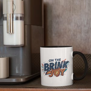 May include: White ceramic mug with a black interior and handle. The mug features the text "ON THE BRINK OF JOY" in a colorful, stylized design. The mug is next to a coffee machine.