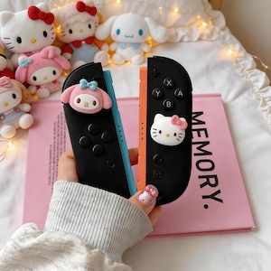 Könnte beinhalten: Schwarze und orangefarbene Nintendo Switch Joy-Con-Controller mit Hello Kitty- und My Melody-Designs. Die Controller sind mit 3D-Charakterfiguren verziert und liegen auf einem rosa Buch mit dem Wort "MEMORY."