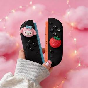 Puede incluir: Mandos Joy-Con negros de Nintendo Switch con diseños decorativos de personajes de dibujos animados y fresas. El mando izquierdo tiene un personaje rosa con un lazo azul. El mando derecho tiene una fresa roja. Los mandos tienen botones negros.