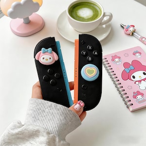 Könnte beinhalten: Schwarze und orangefarbene Nintendo Switch Joy-Con-Controller mit My Melody-Charakterdekorationen. Ein rosa Notizbuch mit My Melody-Design, ein rosa Stift und eine Tasse Matcha sind ebenfalls zu sehen.