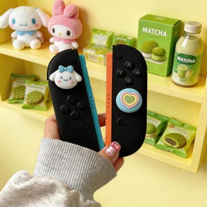 Könnte beinhalten: Schwarze und orangefarbene Nintendo Switch Joy-Con-Controller mit dekorativen Charakterknöpfen. Die Controller werden vor einem gelben Regal mit Matcha-Snacks und Plüschtieren gehalten.