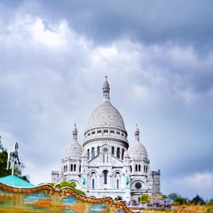 May include: A photograph of the Sacré-Cœur Basilica in Paris, France, under a cloudy sky. The white stone church features multiple domes and towers. A carousel with colorful decorations is visible in the foreground.