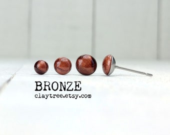 Könnte beinhalten: Vier bronzefarbene Ohrstecker in verschiedenen Größen. Die Ohrringe liegen auf einer weißen Oberfläche. Der Text "BRONZE claytree.etsy.com" befindet sich unter den Ohrringen.