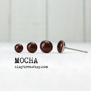 May include: Four brown clay stud earrings of different sizes, with the largest earring shown with a silver post. The earrings are arranged in a line on a white surface. The text "MOCHA claytree.etsy.com" is below the earrings.