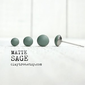 Op de afbeelding: Vier matte saliegroene kleioorbellen in verschillende maten. De oorbellen worden getoond op een wit houten oppervlak. De tekst "MATTE SAGE claytree.etsy.com" is onder de oorbellen afgedrukt.