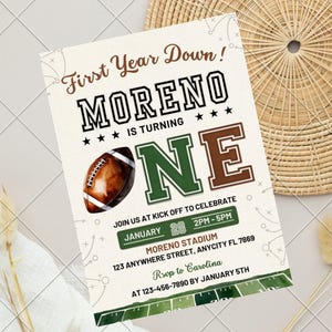 Könnte beinhalten: Eine rechteckige Geburtstagseinladungskarte mit Football-Motiv und dem Text First Year Down! MORENO IS TURNING ONE. Das Design enthält einen braunen Football, grüne und braune Schriftzüge, Veranstaltungsdetails und eine Footballfeld-Grafik.