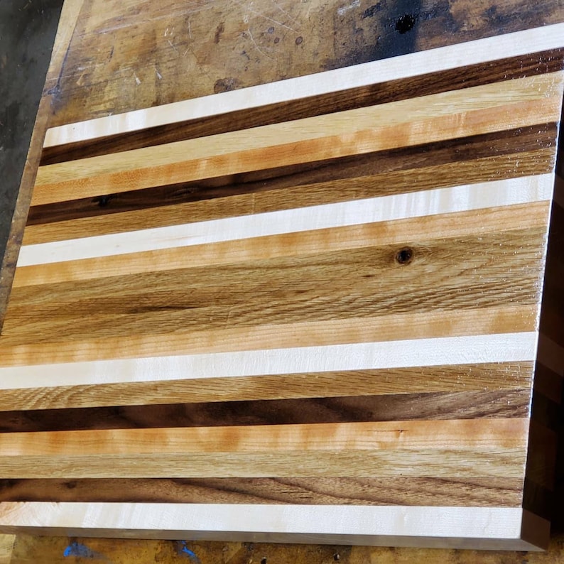 May include: A wooden cutting board with alternating stripes of light and dark wood. The board is made of maple, walnut, and cherry wood.