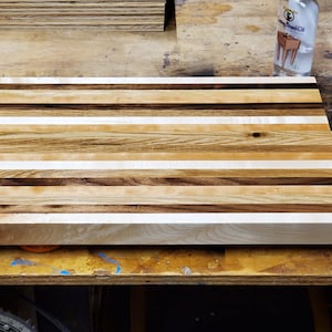 May include: A large, rectangular cutting board made of multiple wood types. The board has a light brown, white, and dark brown striped pattern. The board is sitting on a wooden surface.